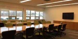 boardroom with large table and green chairs white walls large windows and TV mounted on wall