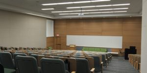 Lecture theater with light brown walls large whiteboards projector and screen and stepped sitting of chairs